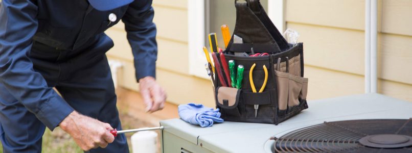 A person in a blue uniform and cap is maintaining an outdoor air conditioning unit with a tool, next to a toolkit on the unit.