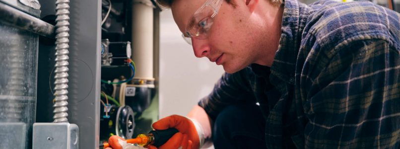 A person in safety glasses and gloves is focused on wiring an electrical component with a screwdriver. They are crouched, working on equipment.