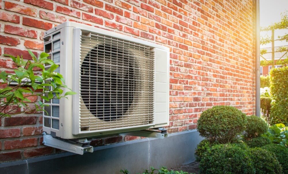 Photo of heat pump on the outside of a house surrounded by bushes.