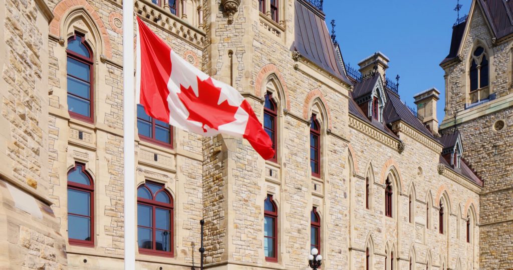 A photo of a Canadian Flag on a pole in front of a parliament building on Ottawa. 