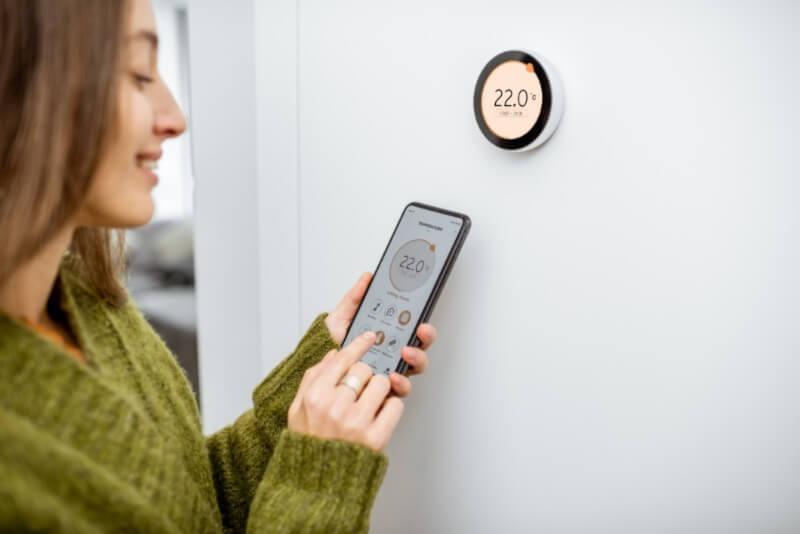 A photo of a woman adjusting a home smart thermostat with a smartphone.