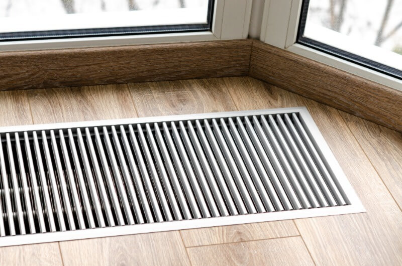 A photo of a hvac vent installed in a wooden floor of a home beside a window.