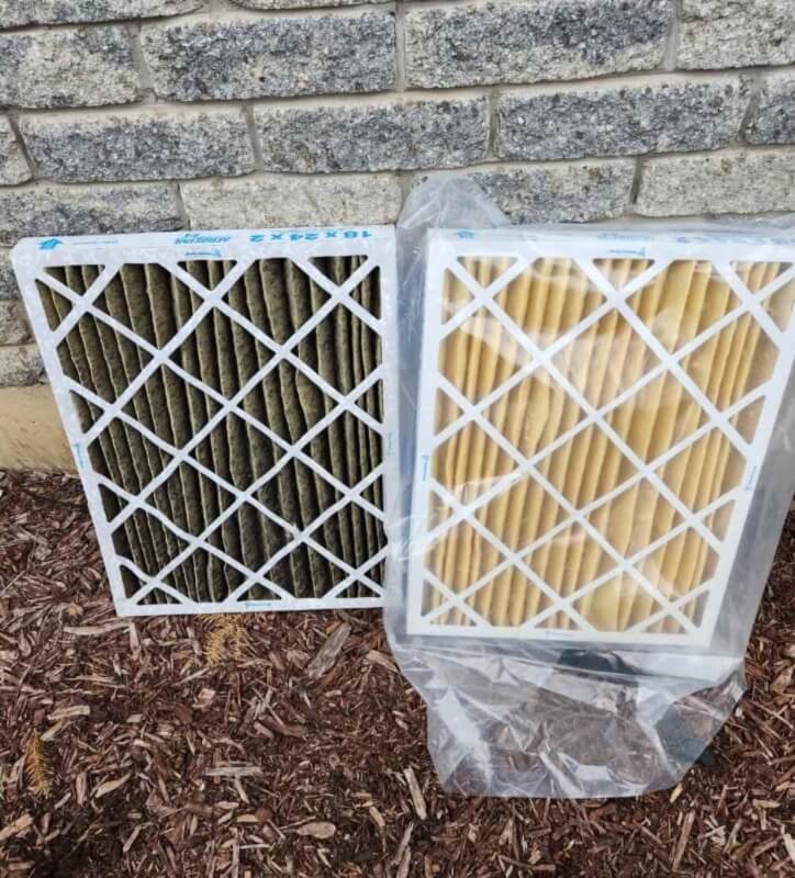 A photo of a dirty furnace filter beside a brand new furnace filter in a bag. They are sitting on the ground beside a house.