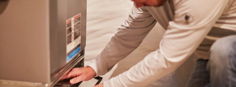 A person is kneeling and working on a household furnace, inspecting or changing the air filter. The setting appears to be a utility room.