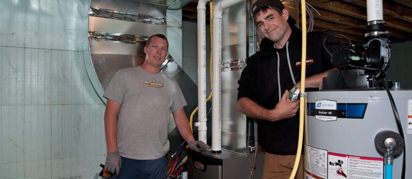 Two people stand in a basement with HVAC equipment. One holds tools; the other leans on a new water heater. They are wearing work attire.
