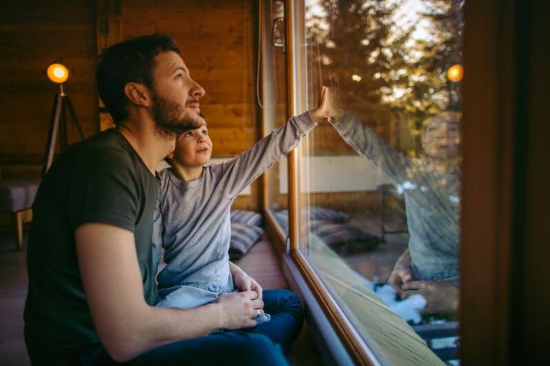 A photo of a man and a child looking out of the window of a log home in the country.