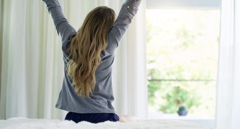 A photo of a woman sitting on the bed, with her arms up in the air stretching . She is facing a window.