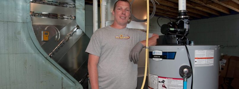 A person in a grey t-shirt and work gloves stands next to a water heater and HVAC system in a utility room, smiling at the camera.