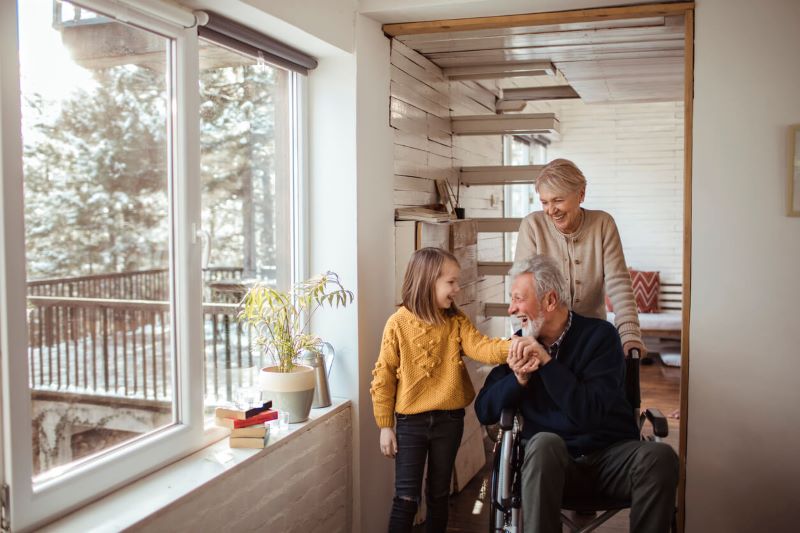 A photo of a girl wearing a yellow sweater standing next to her grandparents in a house. It is winter outside.