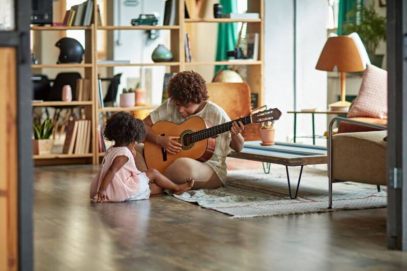 A photo of a mother sitting on the floor of her home playing a guitar, next to her daughter. 