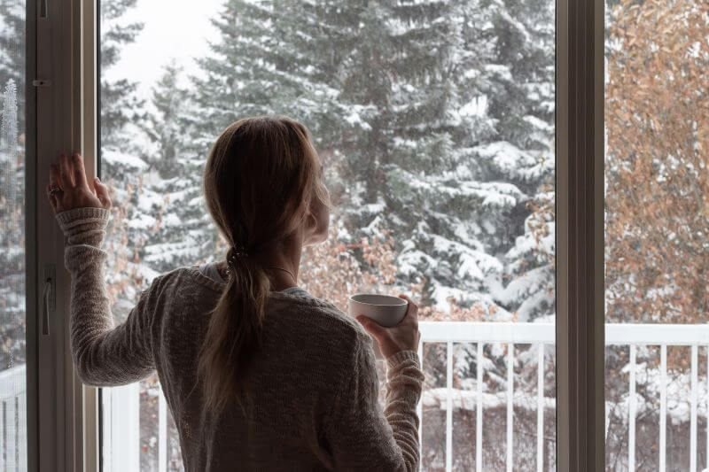 A photo of a woman holding a mug, looking out of a sliding door in her house at trees covered in snow.