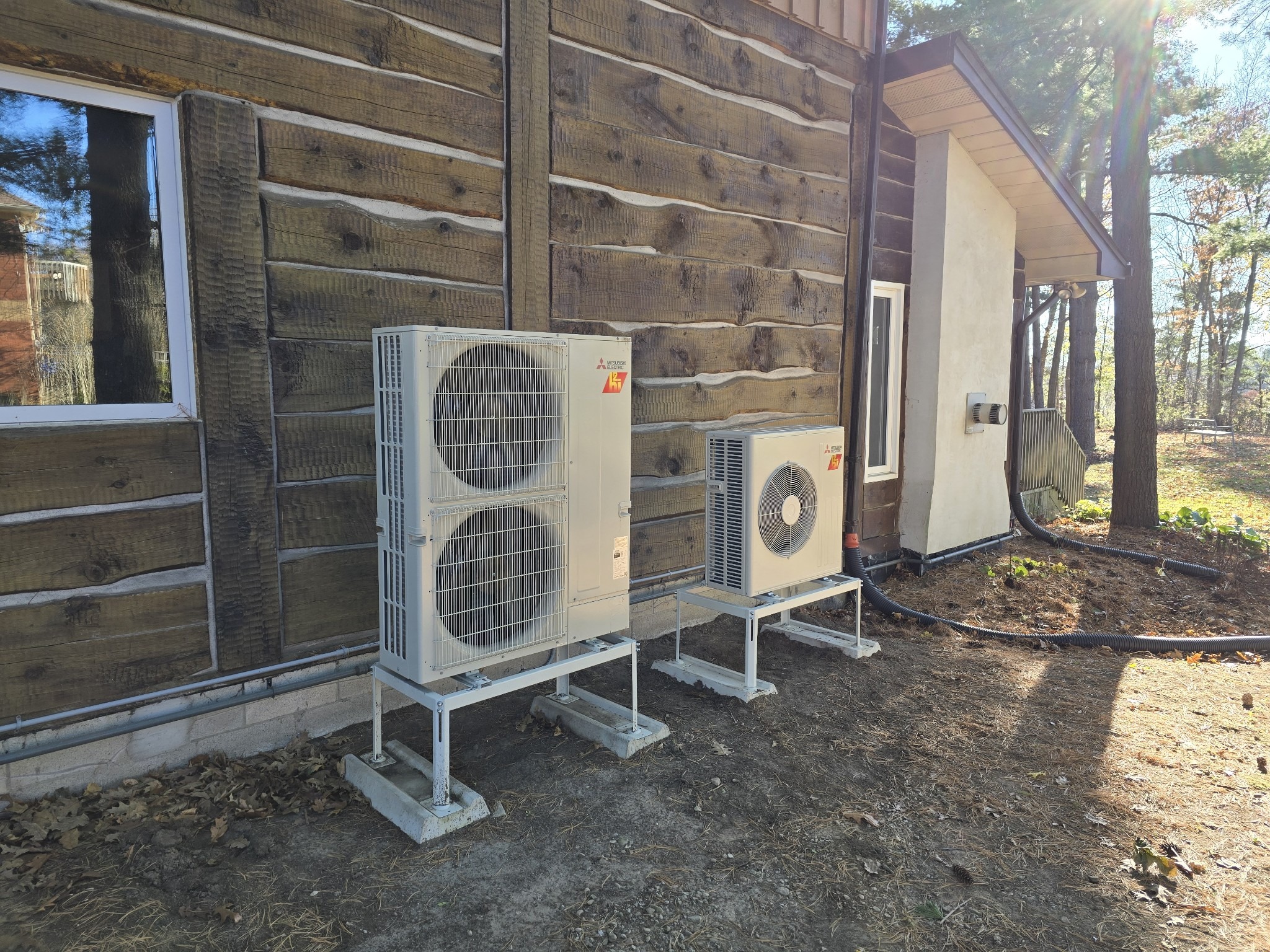 Two outdoor air conditioning units on the side of a wooden building, surrounded by trees and sunlight filtering through branches.