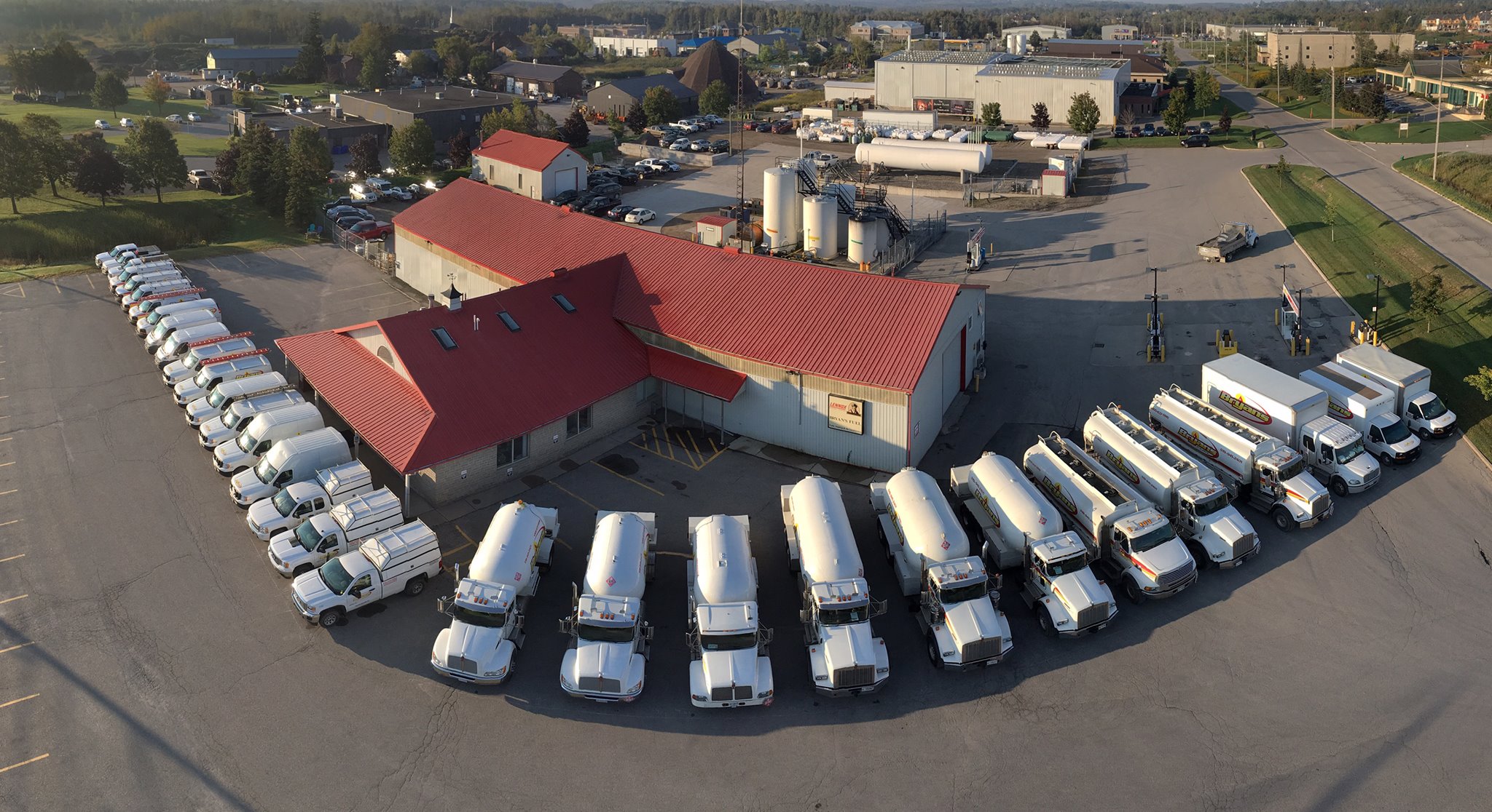 Aerial view of a facility with a red-roofed building, surrounded by numerous white trucks parked in neat rows in a large lot.