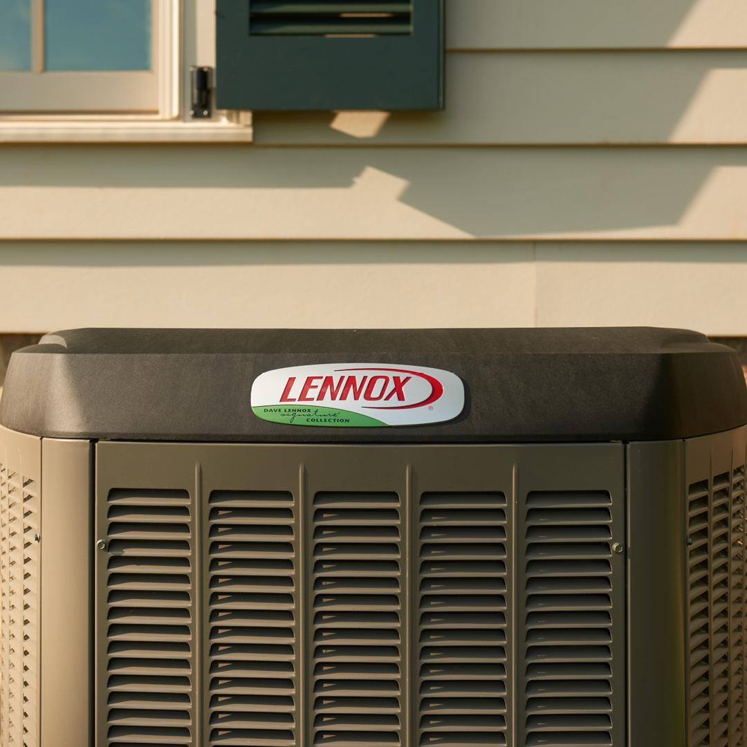 A Lennox air conditioning unit is placed next to a house exterior, featuring beige siding and green shutters, partially shadowed by sunlight.