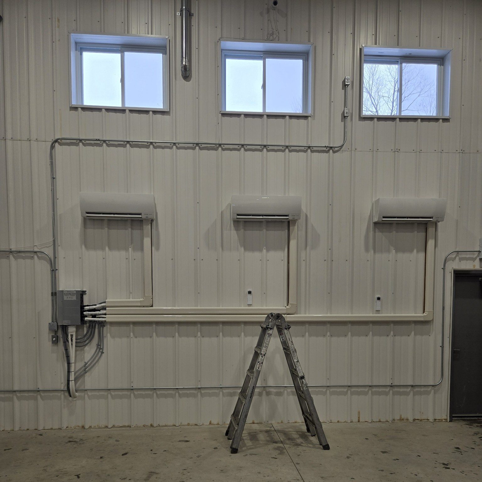 Interior of a warehouse with three air conditioning units, windows, and a metal ladder against a corrugated wall. No people are present.