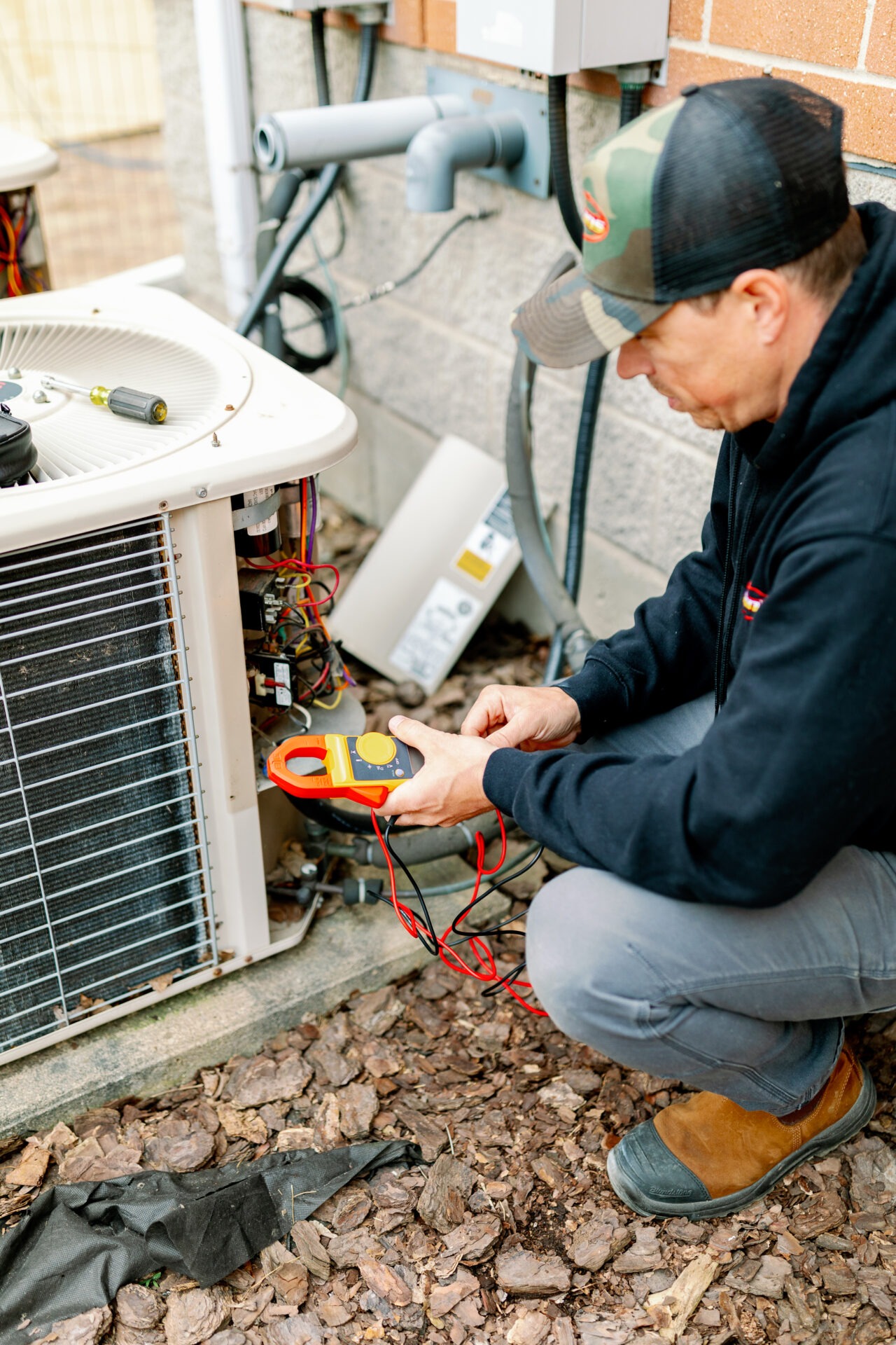 A person squatting next to an air conditioning unit, using a multimeter to check electrical connections, surrounded by mulch and bricks.