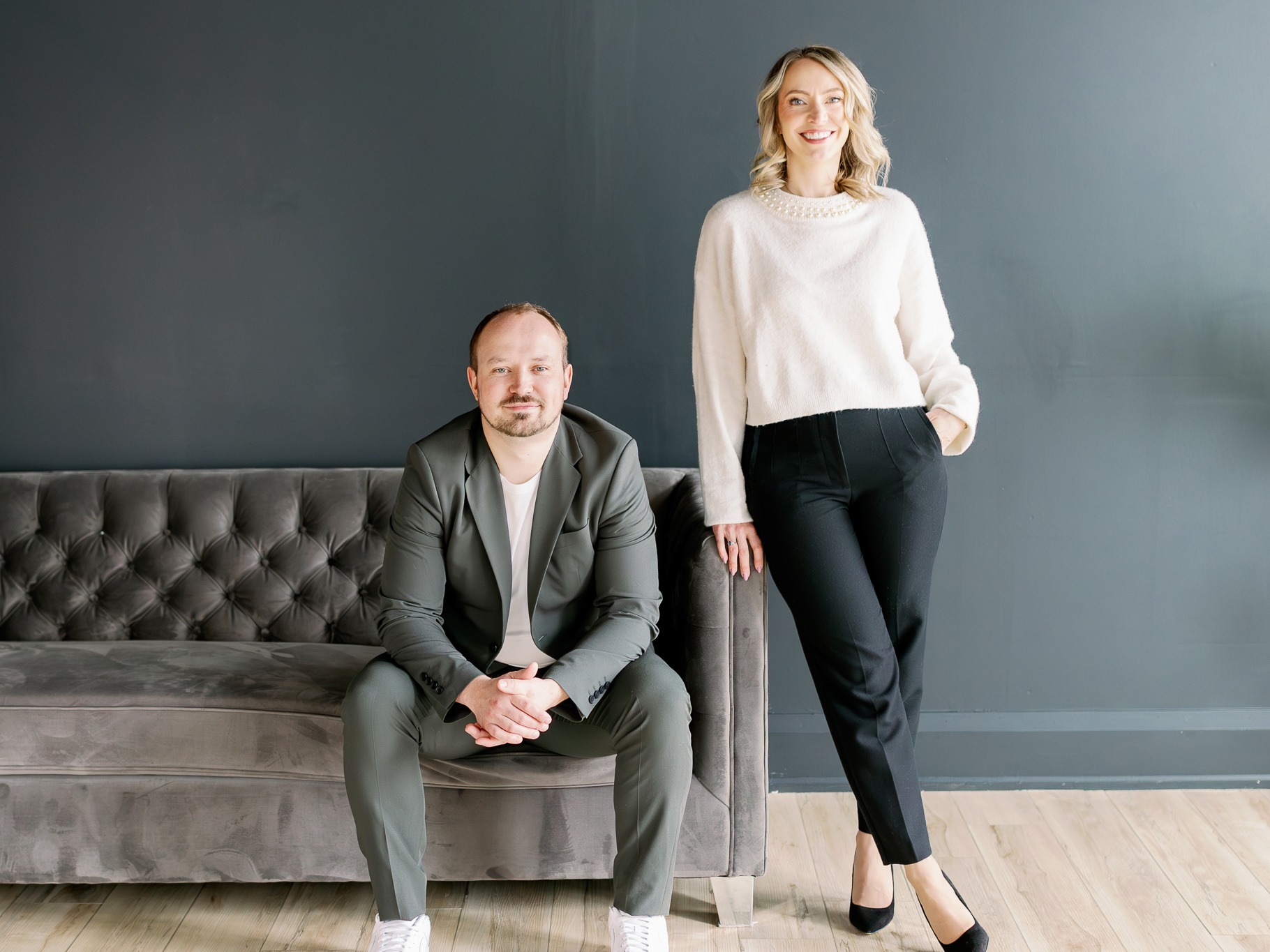 A person seated on a gray couch and another standing beside them, both smiling, against a dark wall in a minimalist setting.