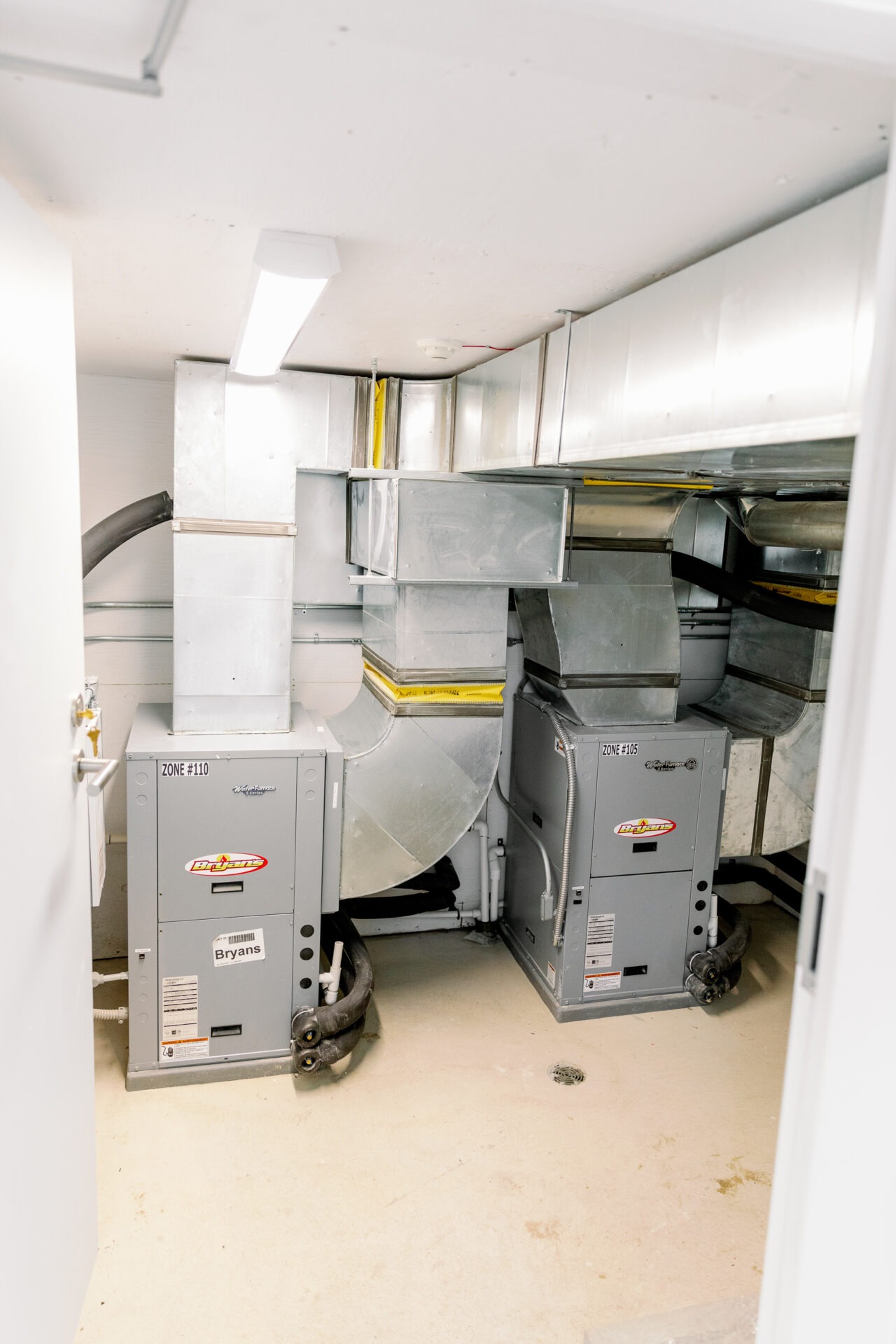 The image shows an indoor HVAC system with ducts and two grey units on a beige floor, under bright fluorescent lighting.