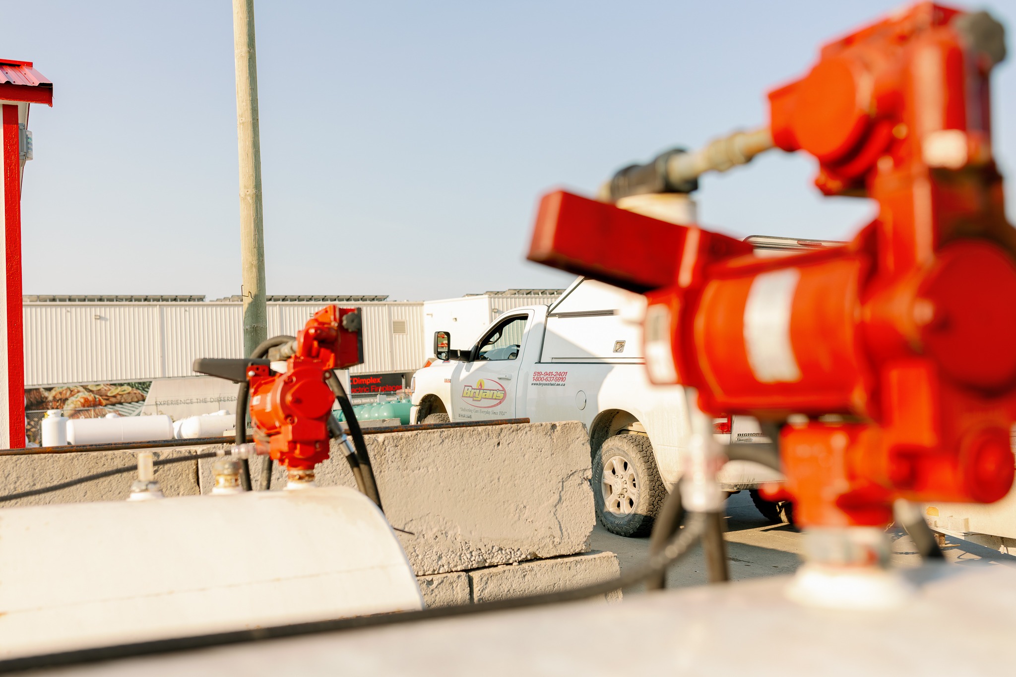 A white truck parked near industrial equipment, with red machinery in the foreground and a large, grey building in the background.