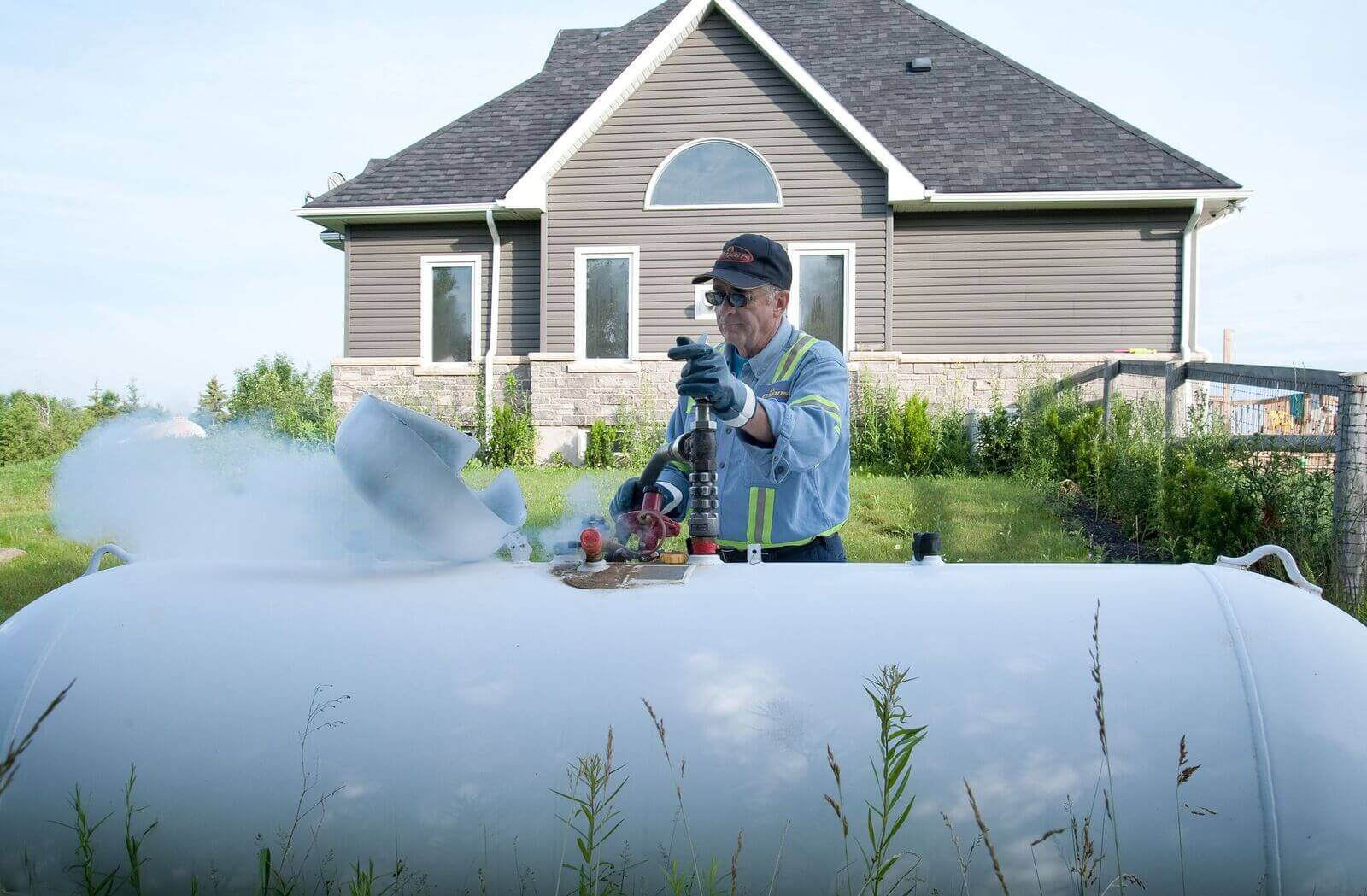 A person works on a large white tank emitting mist, set in a grassy yard with a modern gabled house in the background.