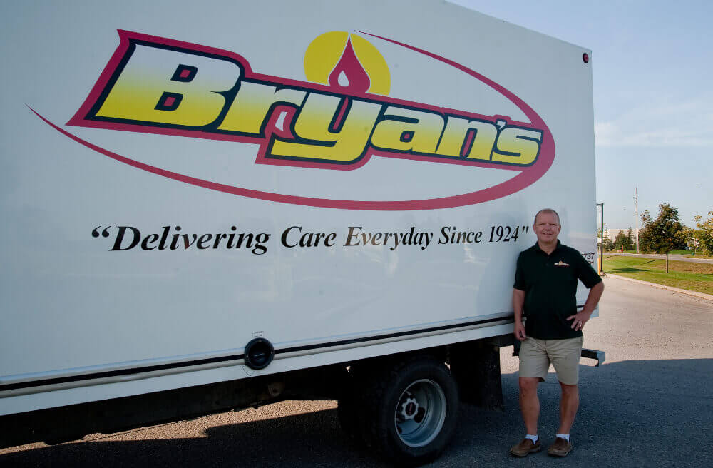 A person stands beside a large truck with "Bryan's" logo, emphasizing care delivery since 1924, on a sunny day outdoors.