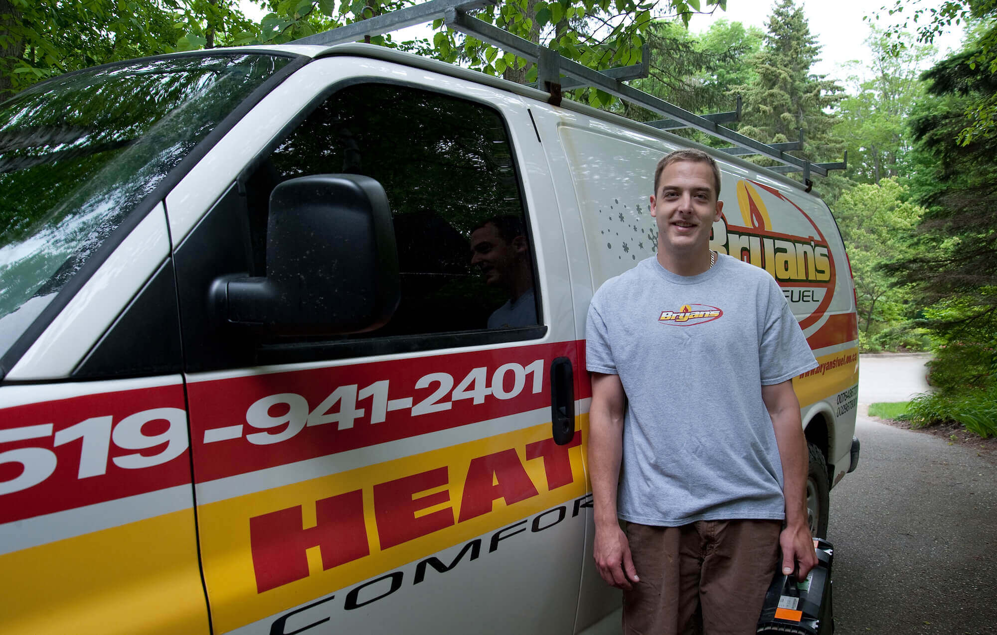 A person stands beside a branded service van on a wooded driveway, wearing a gray t-shirt and holding a tool case.