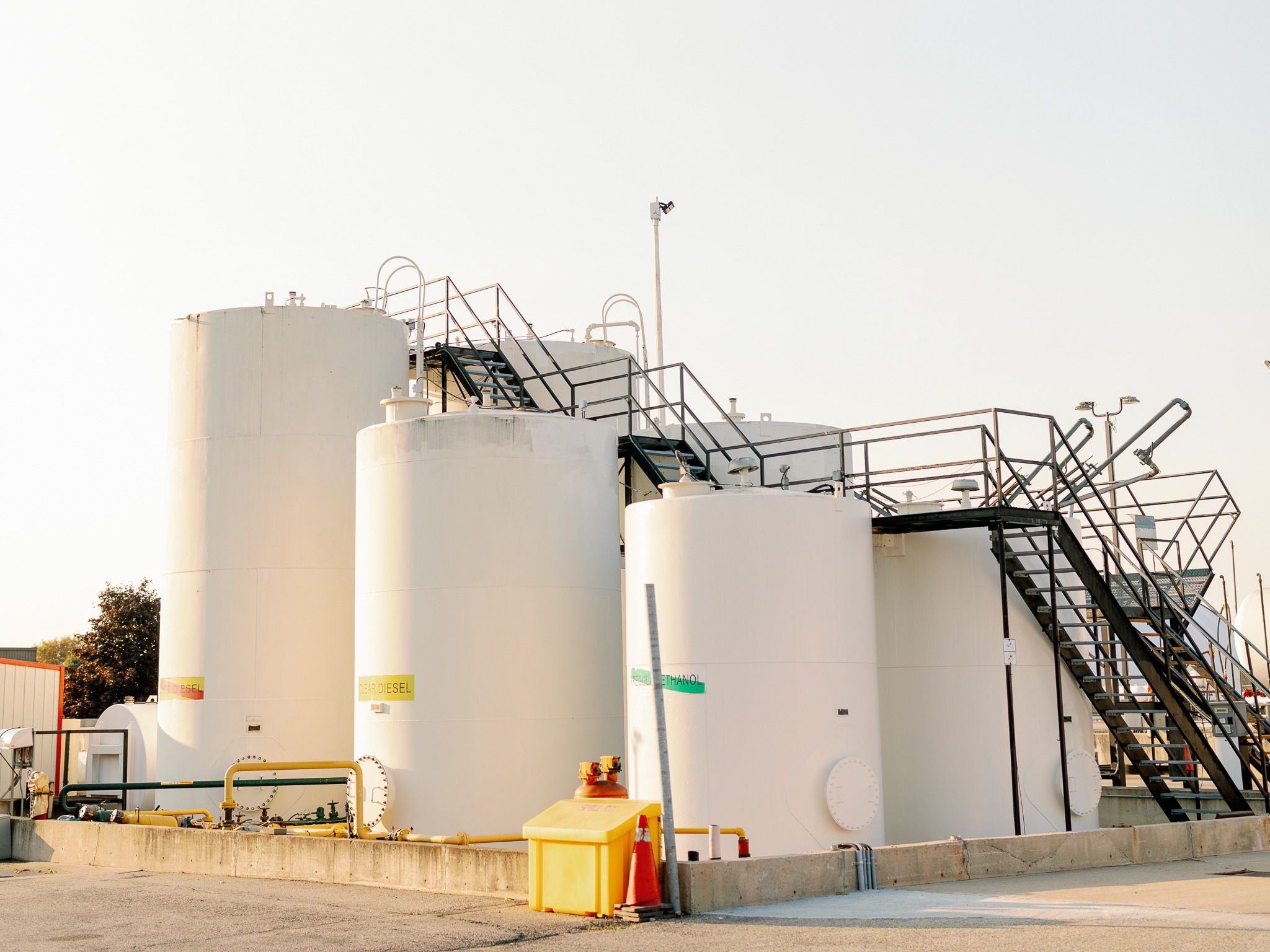 Industrial site with large white tanks labeled biodiesel and ethanol, surrounded by pipes, ladders, and safety equipment under a clear sky.