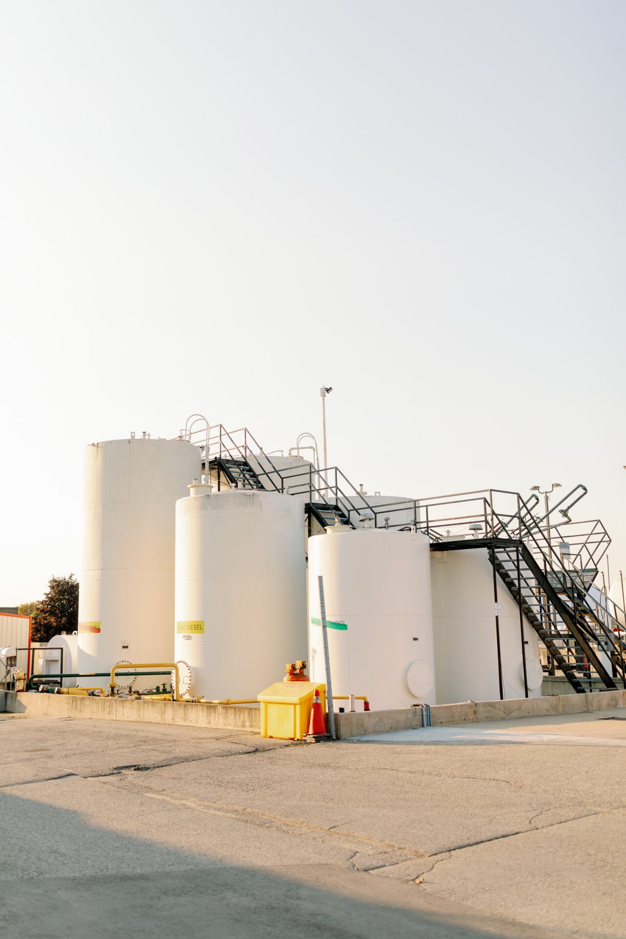 Industrial site with large white storage tanks, labeled biodiesel and ethanol. Metal staircases connect tanks; equipment and traffic cones visible on concrete surface.