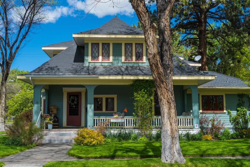 Classic two-story craftsman house with green siding, surrounded by trees and landscaped garden, set against a clear blue sky and lush lawn.