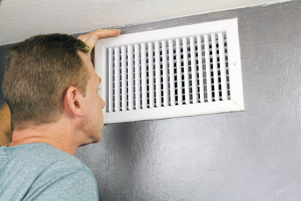 A person inspects a white air vent on a gray wall, examining it closely. The image captures a household maintenance scene.