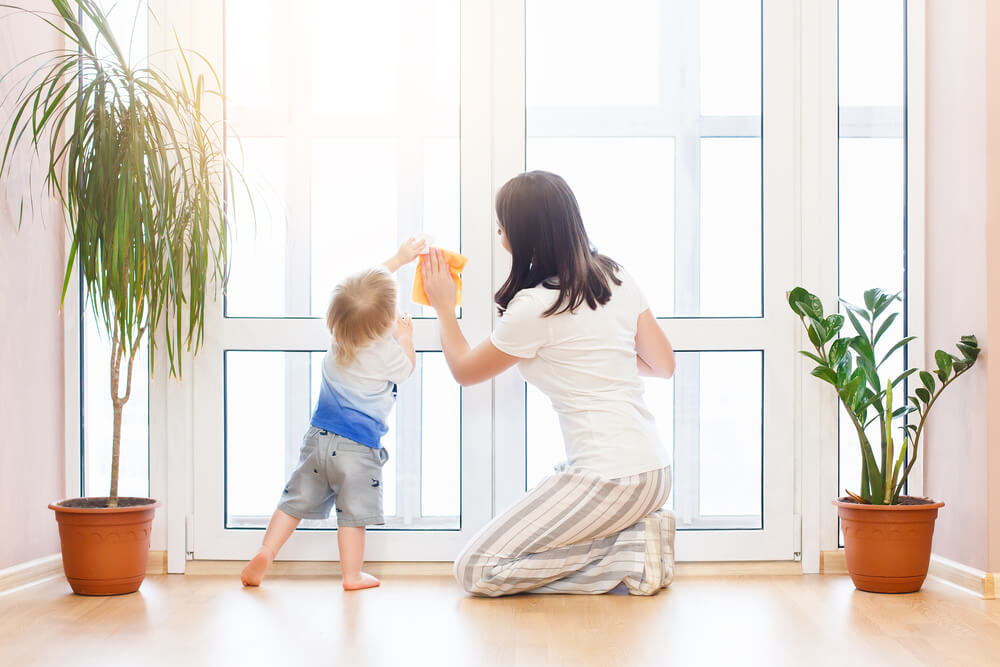 A person and child in a bright room clean a large window. Two potted plants decorate the wooden floor.