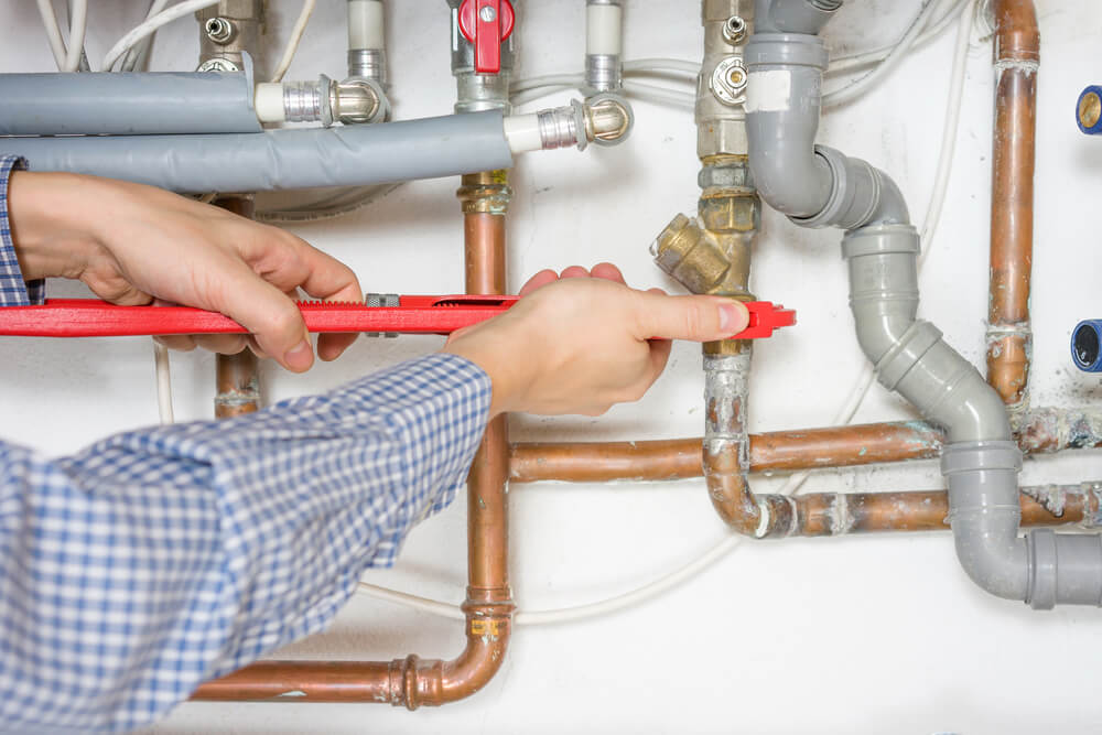 A person uses a red wrench to adjust pipes and valves on an intricate plumbing system mounted on a white wall.