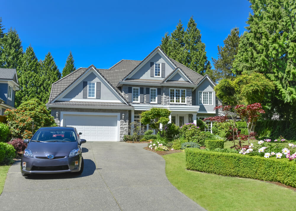 A modern, gray suburban house with manicured garden and parked car on driveway under a clear blue sky, surrounded by lush trees.