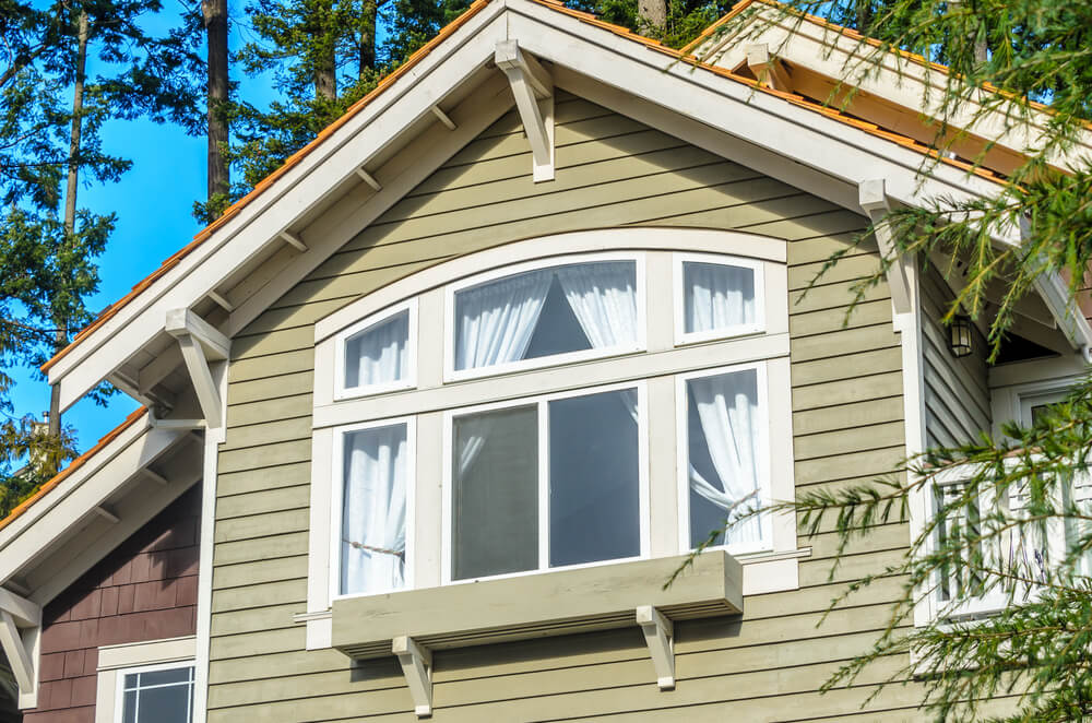 A close-up of a house's upper facade with large windows and white curtains, surrounded by tall green trees under a clear blue sky.