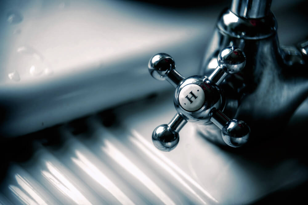 Close-up of a vintage-style metal faucet handle marked "H" for hot, set against a white porcelain background with water droplets.