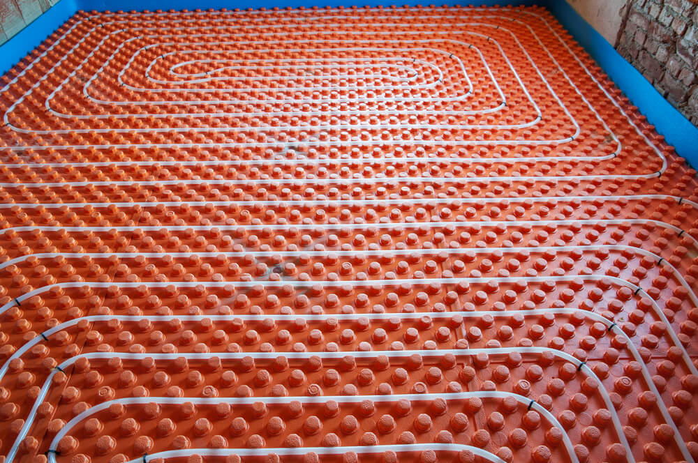 Orange underfloor heating system installation with a grid of white pipes on a textured surface, surrounded by a blue frame and brick walls.