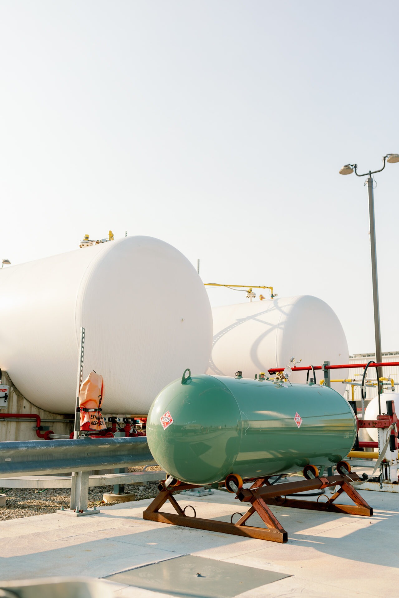 Industrial site with large white storage tanks and a smaller green tank. Pipes, valves, and safety equipment are surrounding the installation.