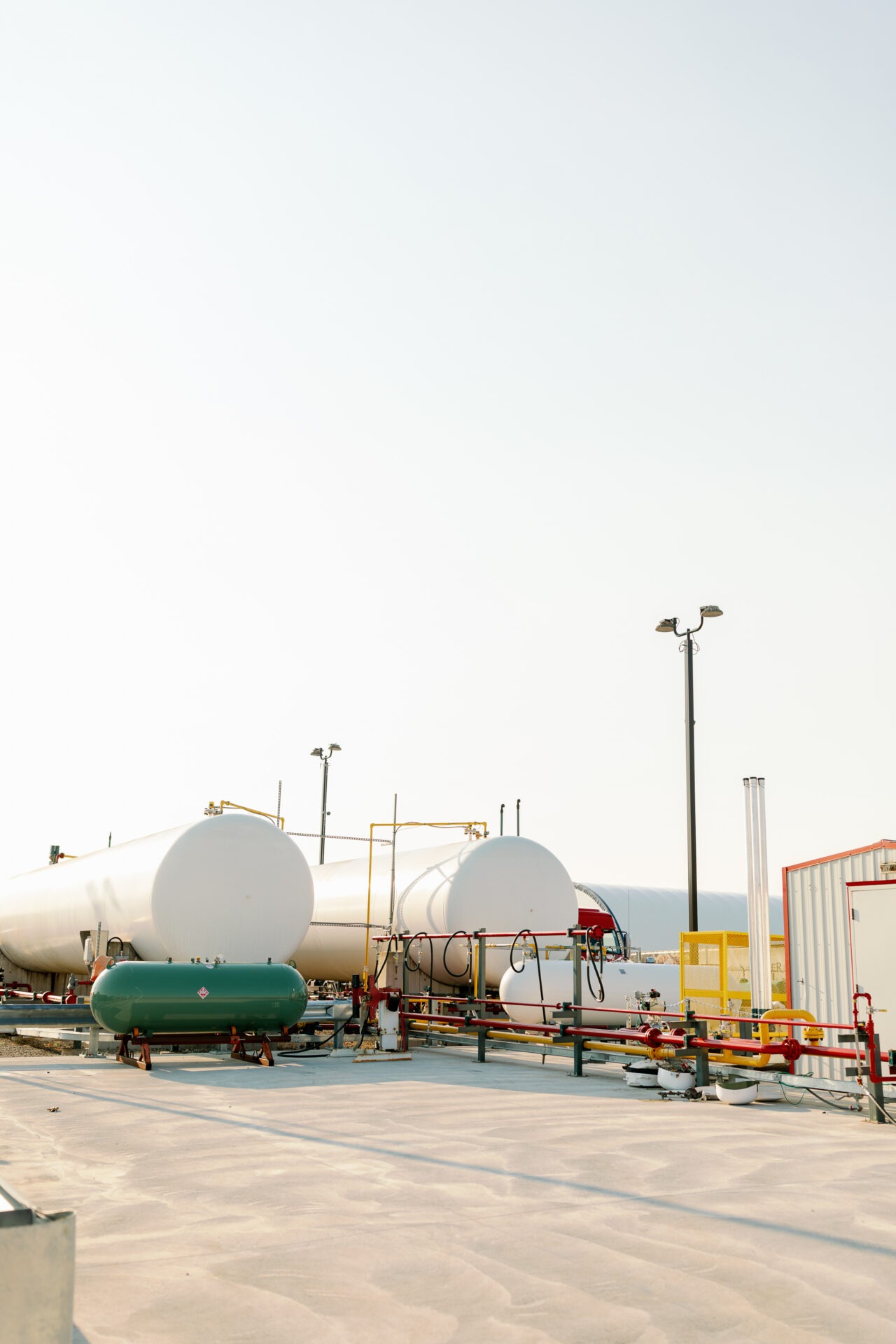 Several large industrial tanks and pipes are set in a bright open area, with clear skies and safety equipment visible.