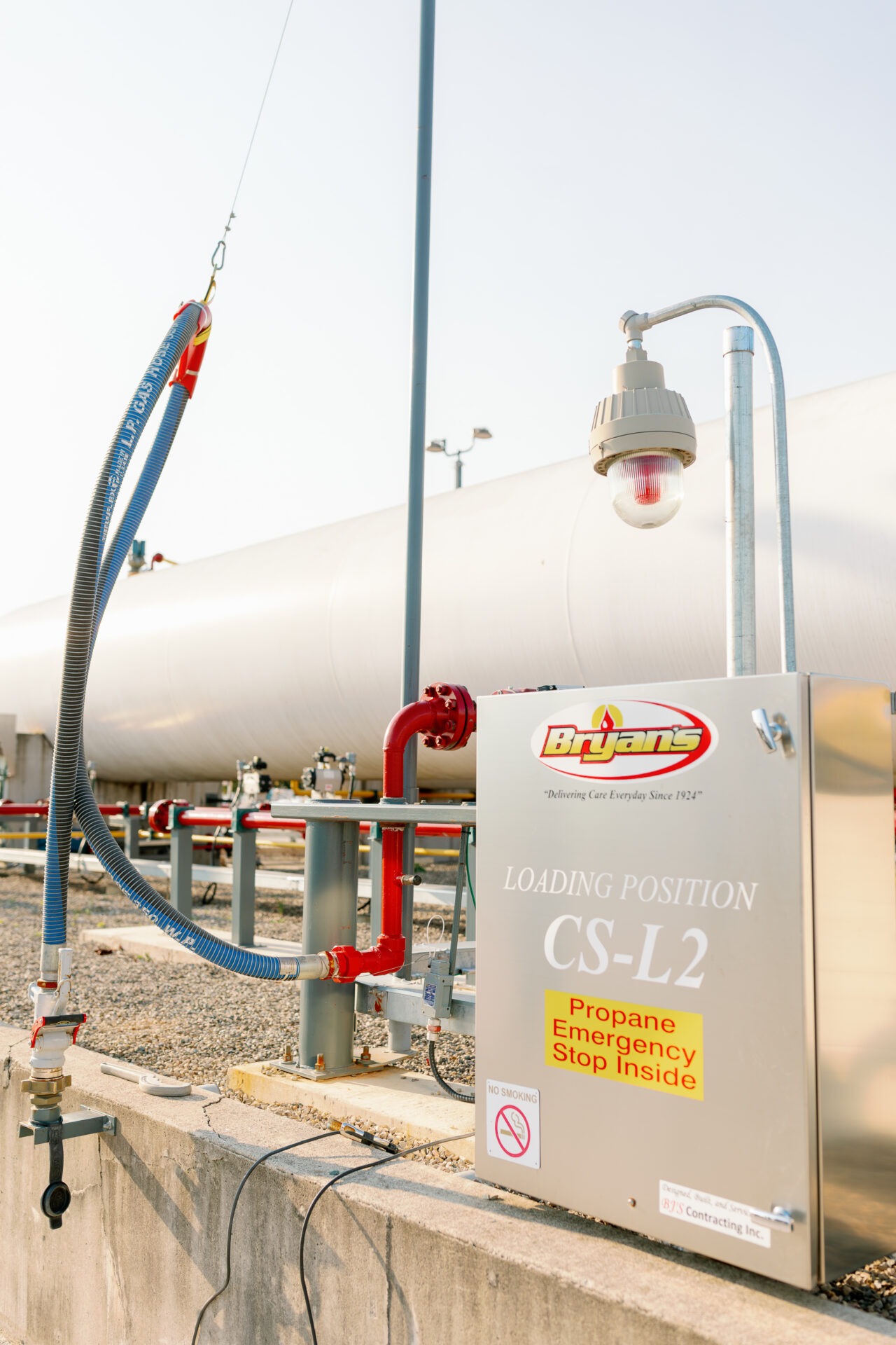 Industrial propane loading station with safety signage and hoses. A metal enclosure labeled "Bryan's" and emergency stop information is prominently displayed outdoors.