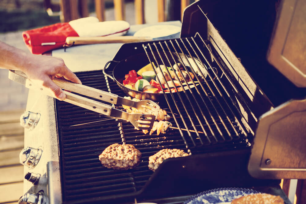 A person is grilling burgers and vegetables on an outdoor barbecue, using tongs. A red oven mitt is nearby.