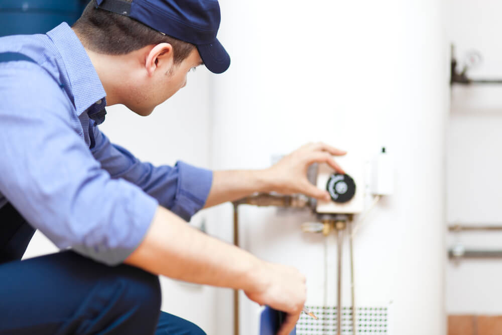 A person in a blue shirt and cap adjusts a temperature dial on a white water heater, focusing intently on the task.
