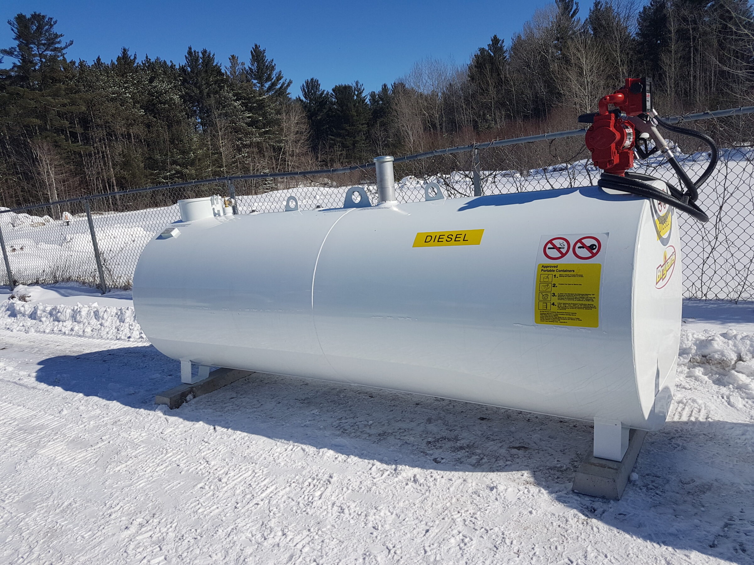 A white diesel fuel tank with red pump sits in snowy, fenced area surrounded by trees under clear blue sky.