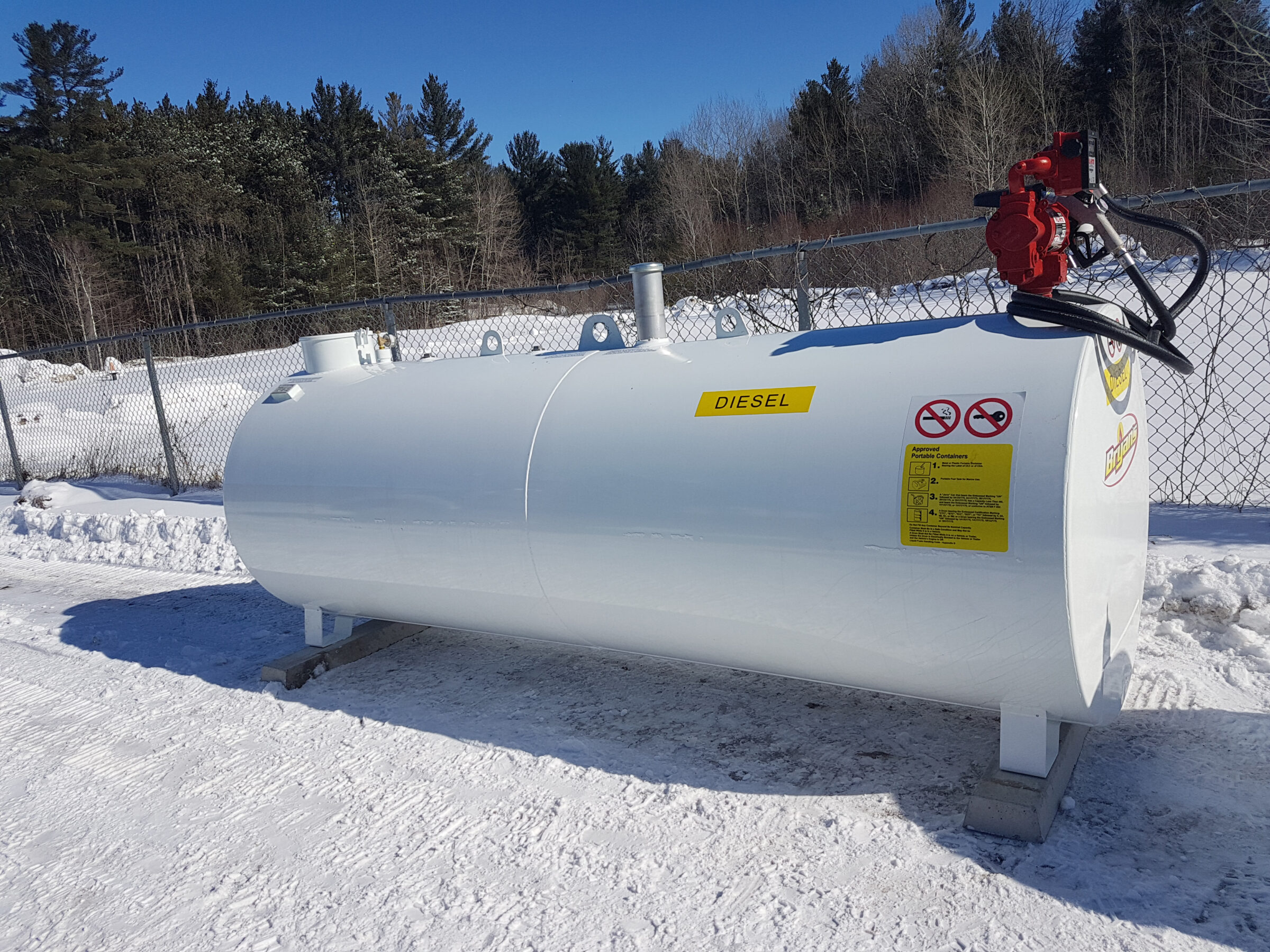 A white diesel tank with red pump sits in a snowy area, surrounded by a chain-link fence and pine trees under a clear blue sky.