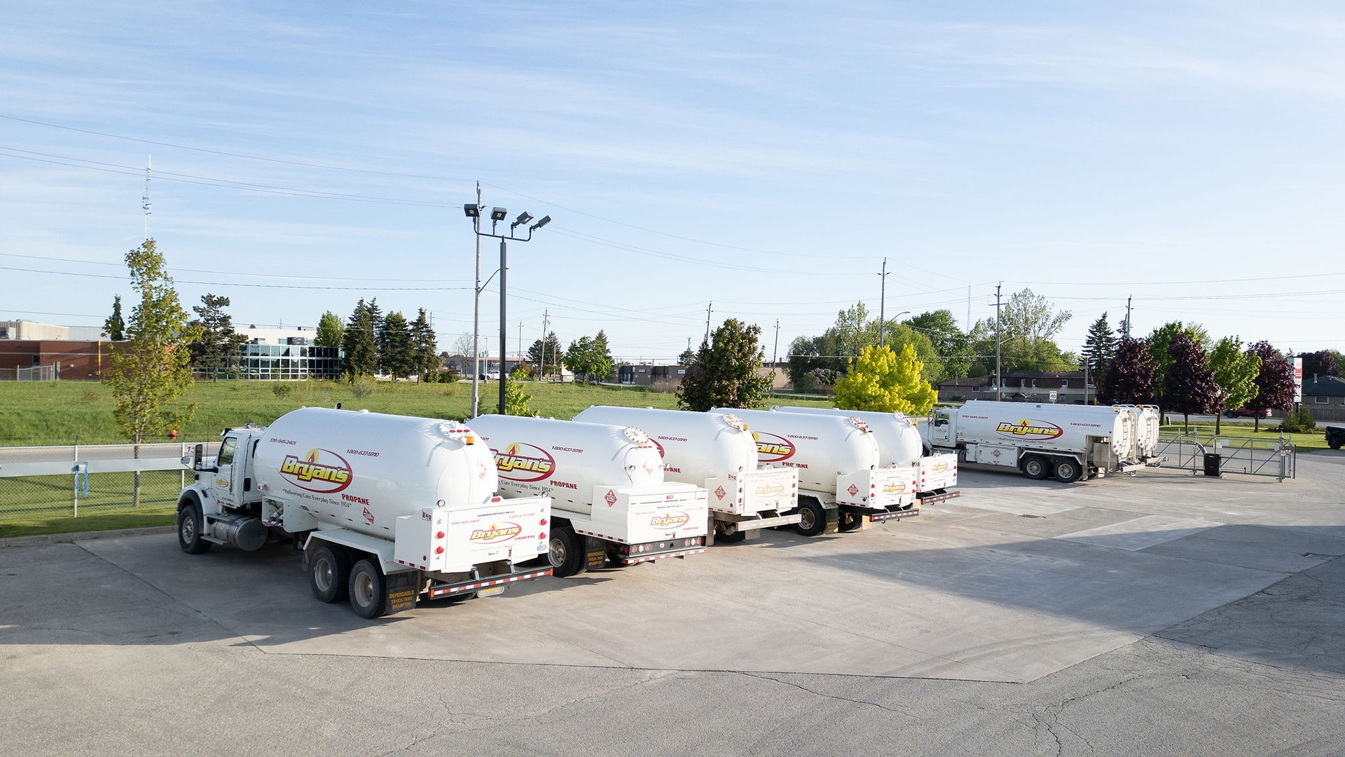 Six propane trucks parked in a lot surrounded by greenery and buildings under a clear blue sky, with no people present.