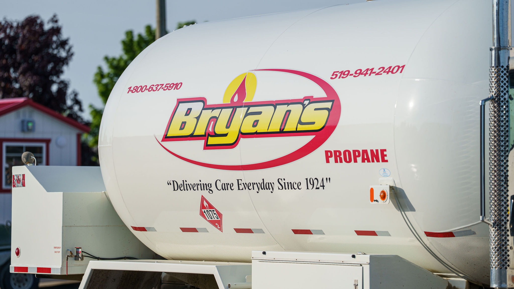 A white propane truck with "Bryan's Propane" branding, parked near a small building, trees visible in the background, sunny day.