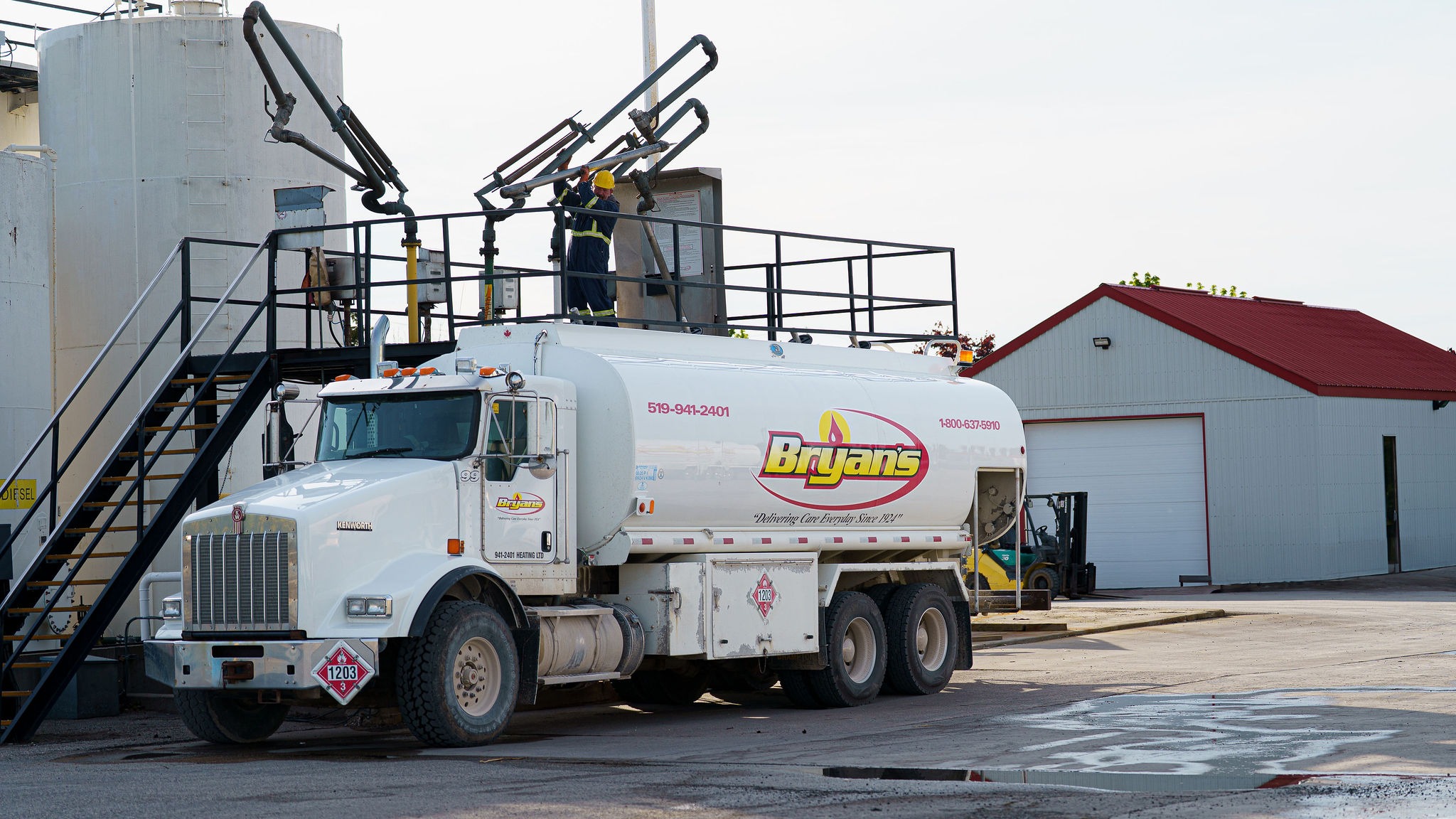 A person in a hard hat operates equipment atop a fuel truck labeled "Bryan’s" near a small industrial building and storage tanks.