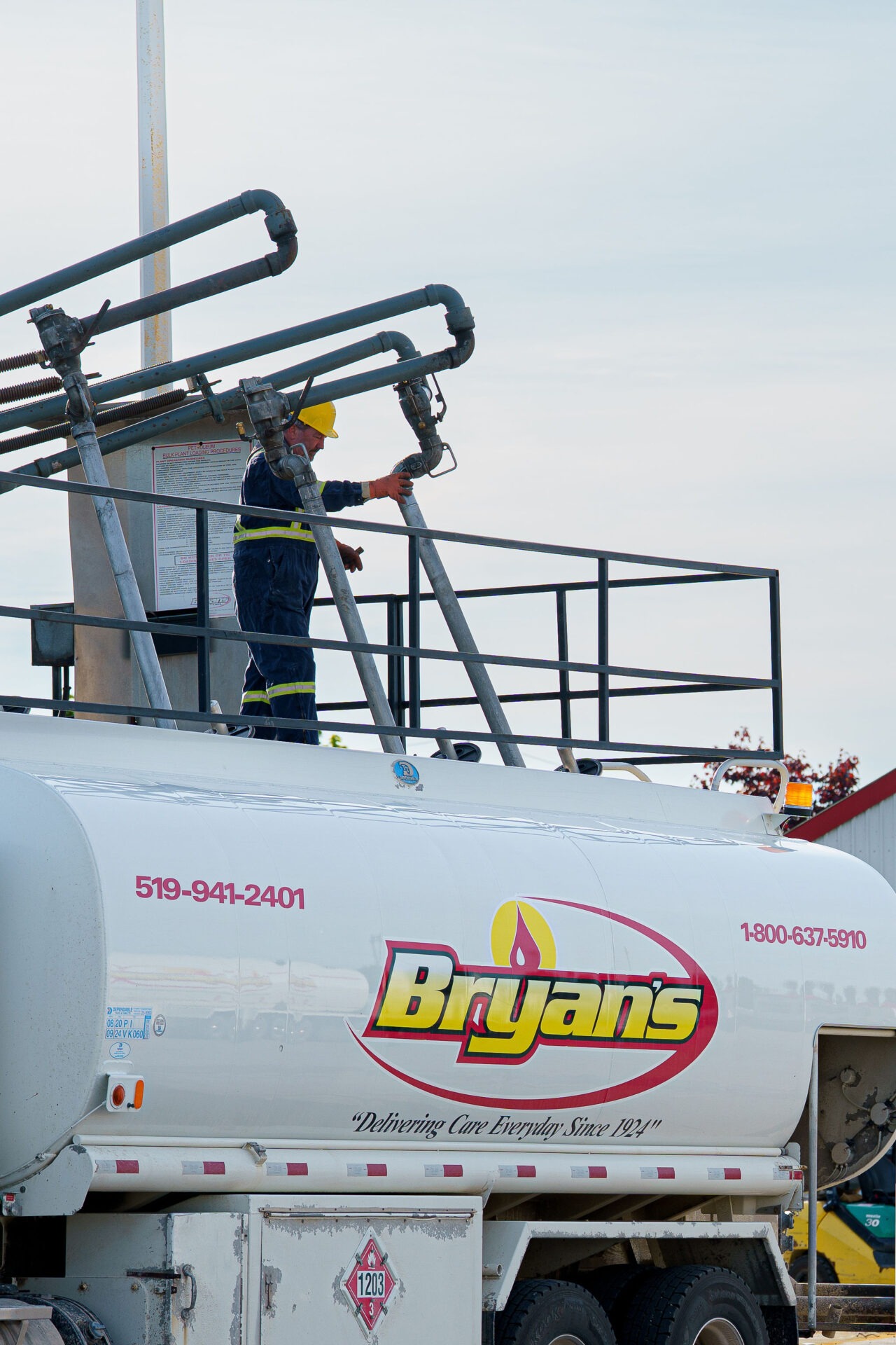 A person in safety gear works atop a white tanker truck labeled "Bryan's," surrounded by industrial piping against a clear sky.