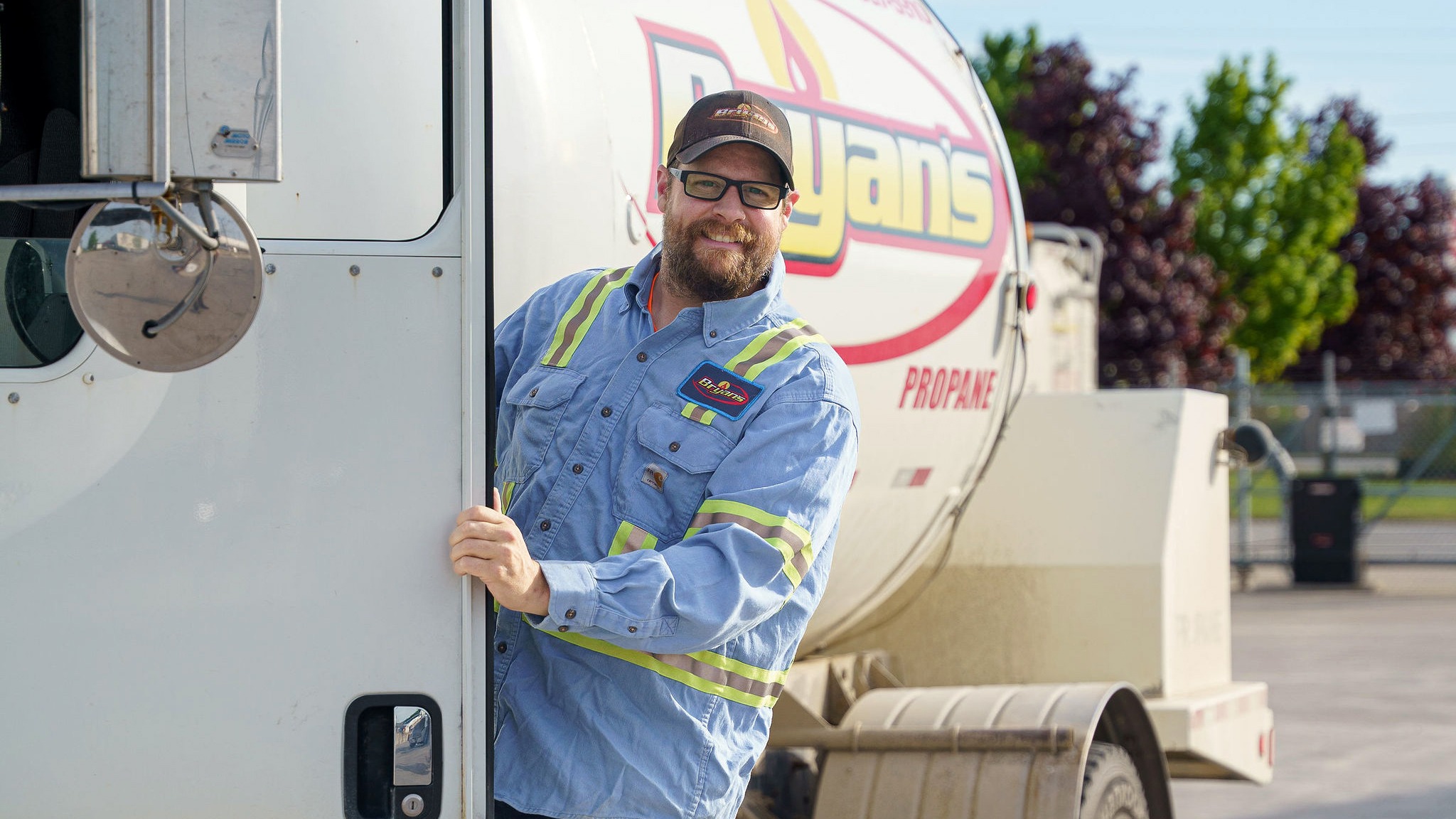 A person with glasses, wearing a uniform, standing by a propane truck, smiling. Trees and blue sky in the background.