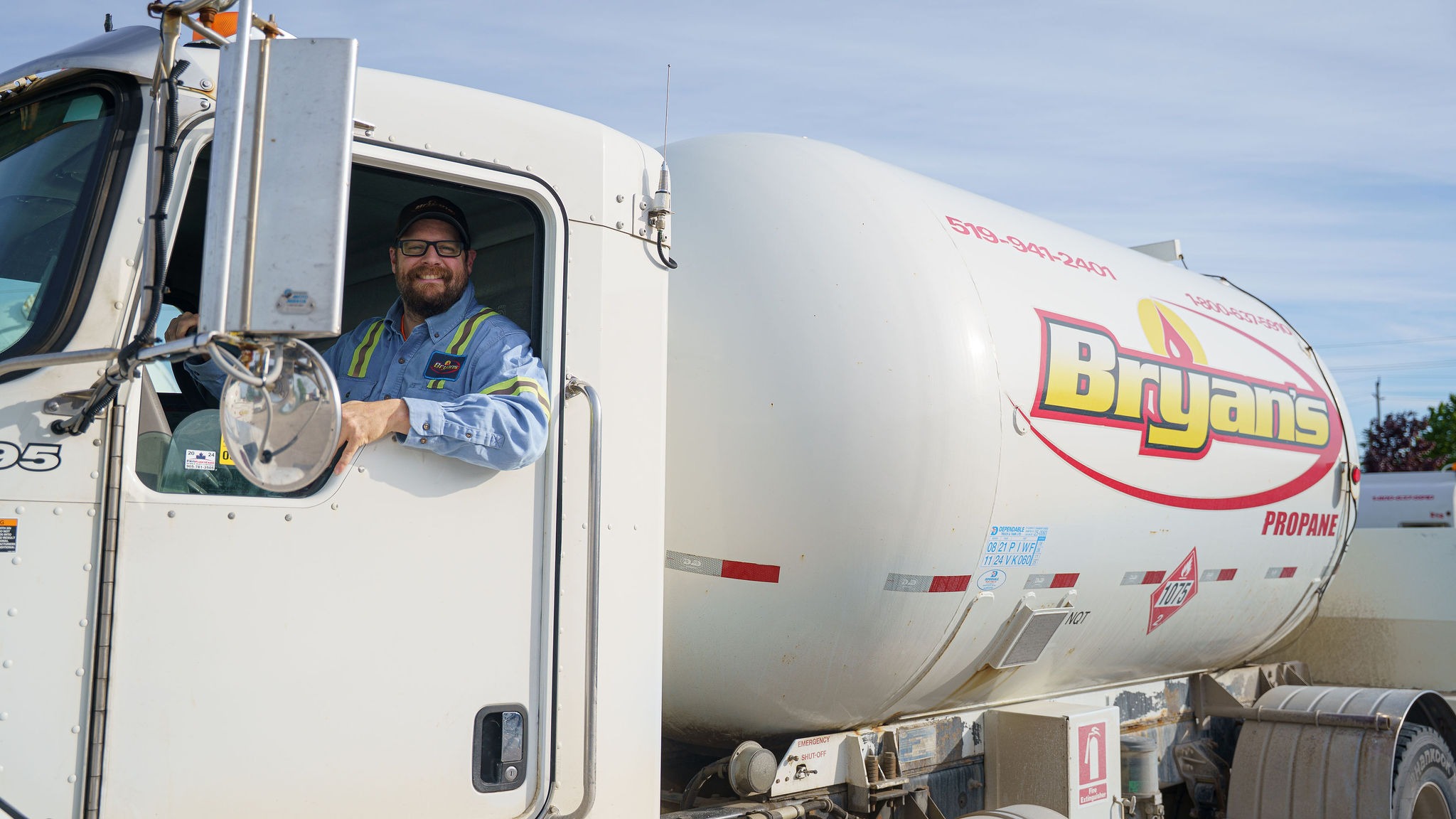 A person smiles from a parked propane truck labeled "Bryan's" under a clear blue sky, wearing safety gear and glasses.