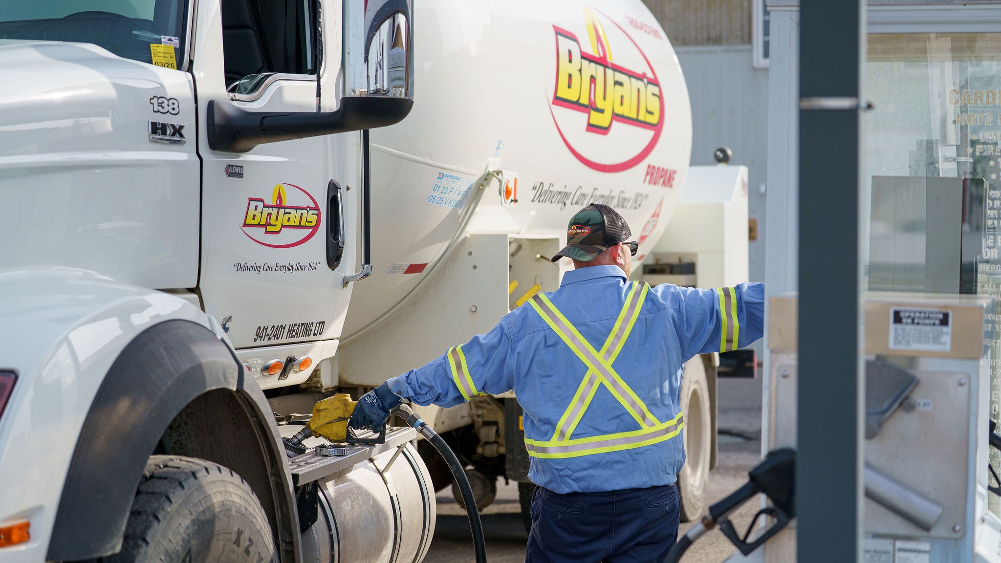 A person in a safety vest refuels a Bryan's propane truck at a gas station, managing the hose and equipment.